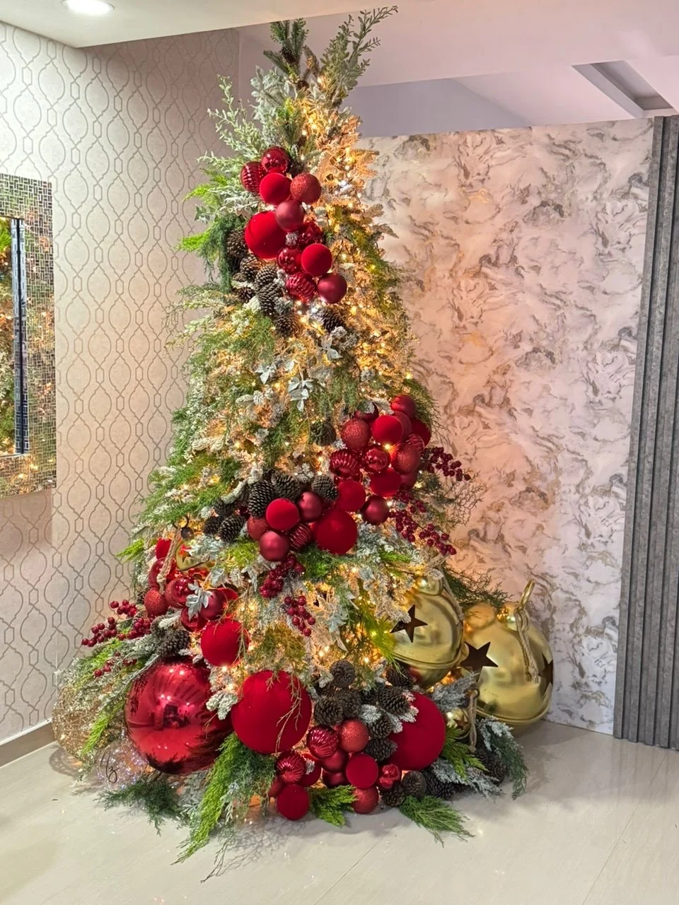 Christmas tree decorated with oversized red baubles, pinecones, mixed greenery and warm lights beside gold bell decorations.