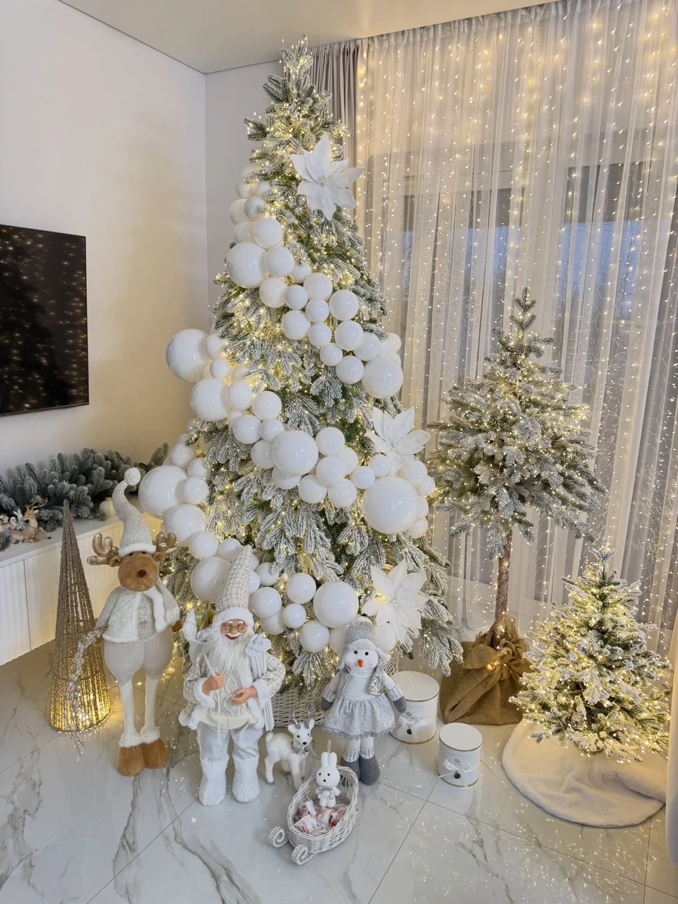 Three illuminated flocked Christmas trees with white baubles, white flowers and festive figures against curtain lights.
