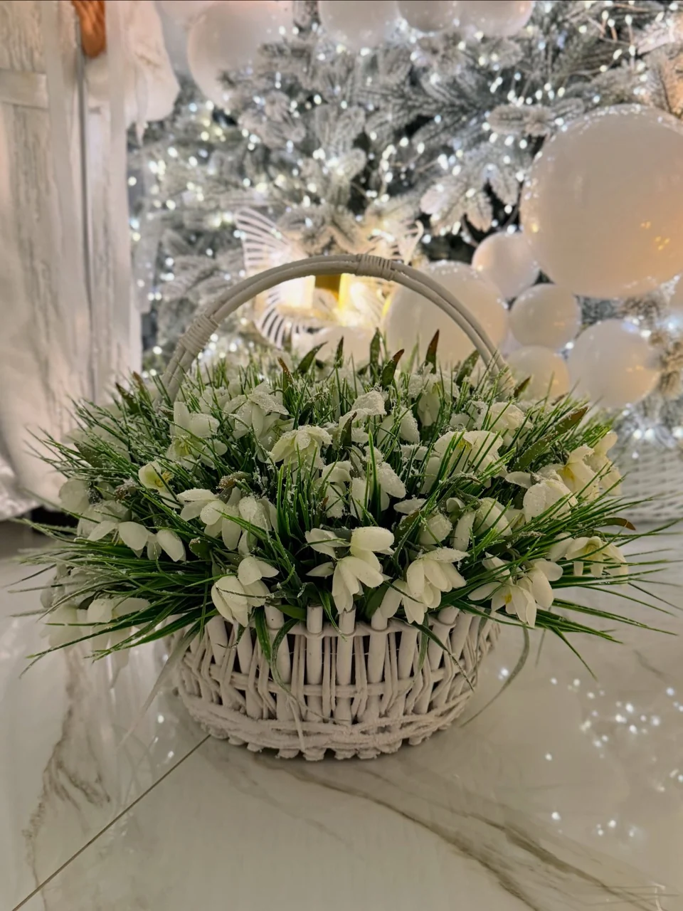 White flower stems and greenery in a white wicker basket in front of a softly lit Christmas tree.
