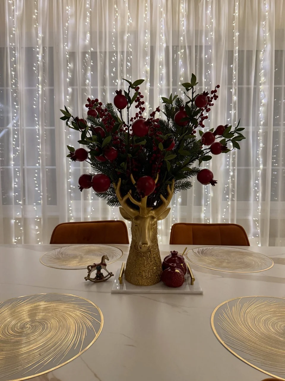 Red fruit and greenery arranged in a gold deer-shaped vase on a dining table in front of curtain lights.