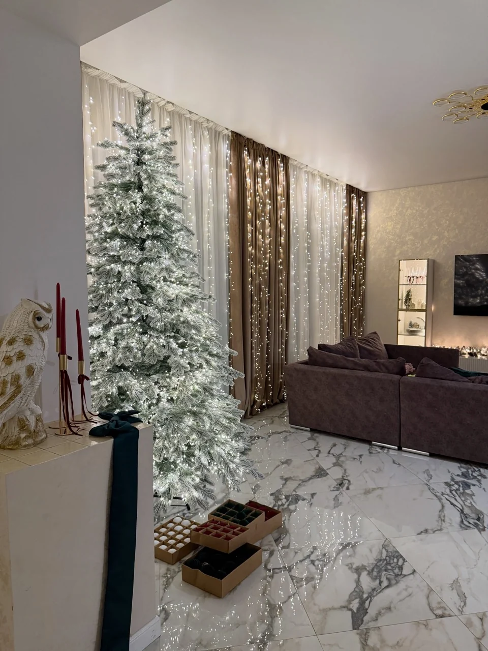 Flocked Christmas tree and curtain lights beside a sofa, with ornament boxes at the tree base.