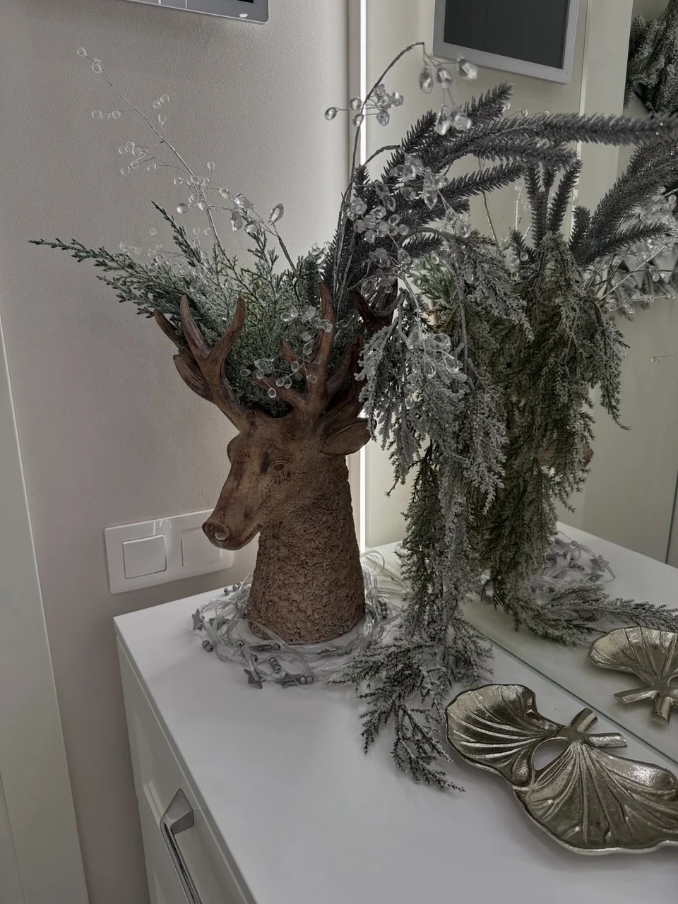 Deer-head vase filled with frosted greenery and clear decorative stems on a white console beside a mirror.