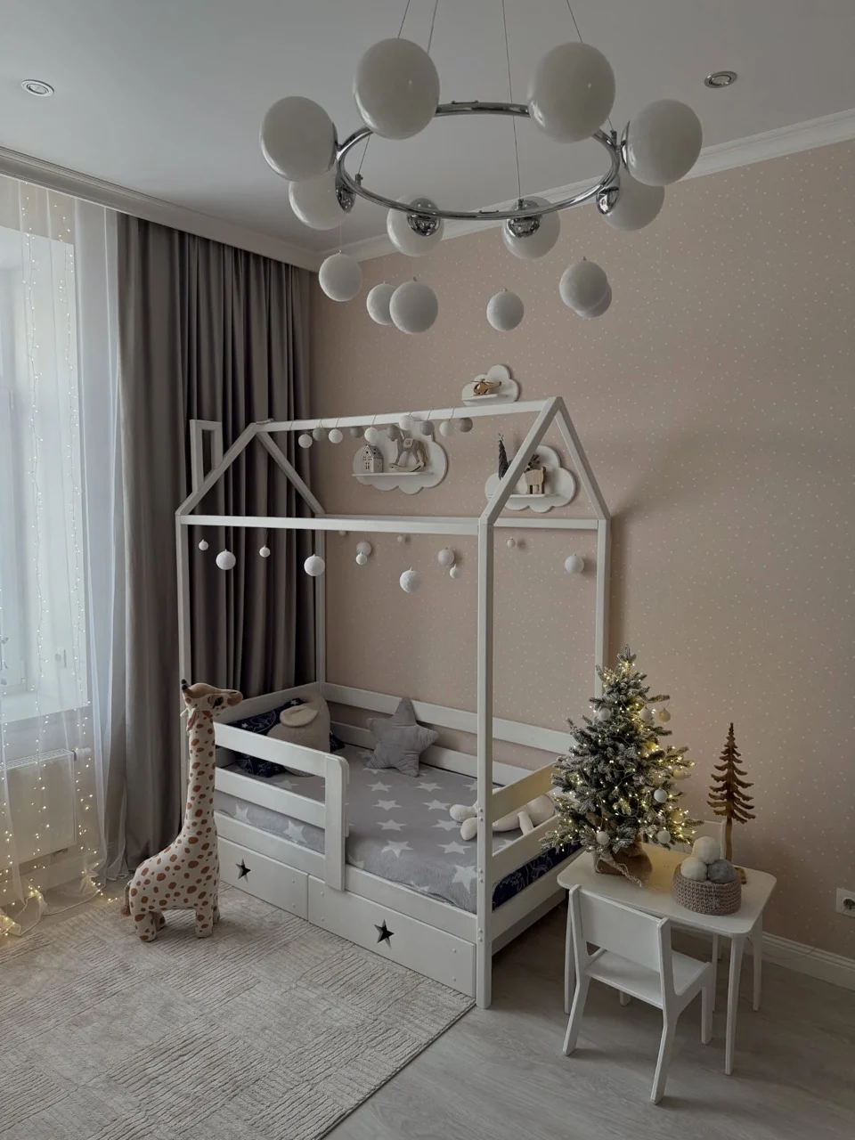 Small illuminated Christmas tree beside a white house-frame bed with a white bauble garland and giraffe toy.