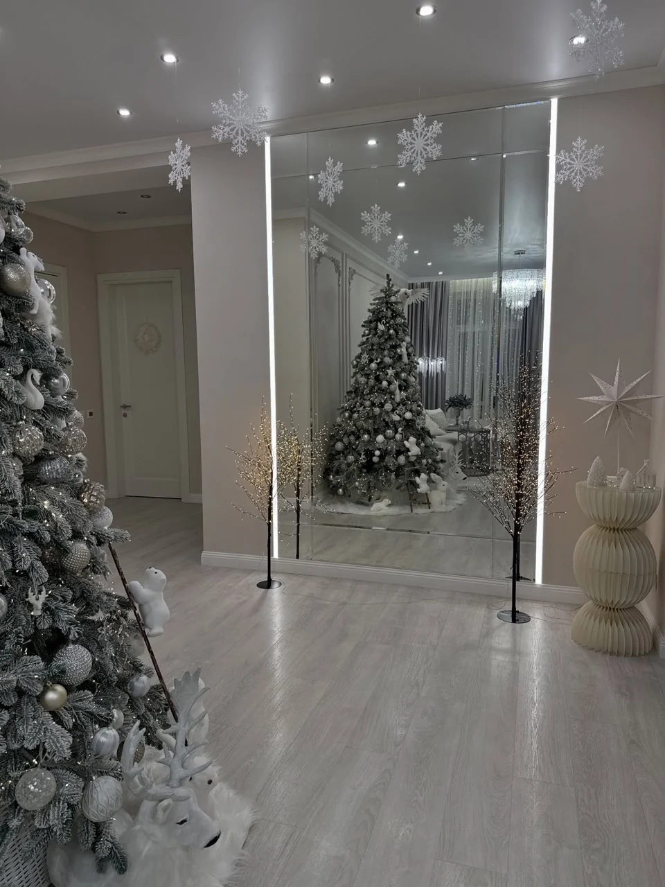 Decorated woodland Christmas tree reflected in a tall mirror beneath hanging snowflakes, with illuminated twig trees beside it.