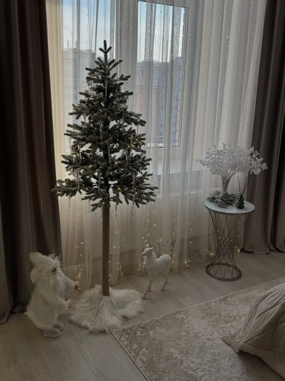 Slender Christmas tree on a wrapped trunk beside white winter figures and curtain lights in a bedroom.