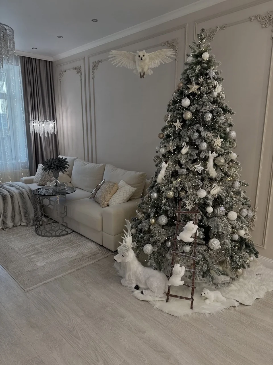 Silver and white Christmas tree with woodland animals, a small wooden ladder and a suspended owl beside a cream sofa.