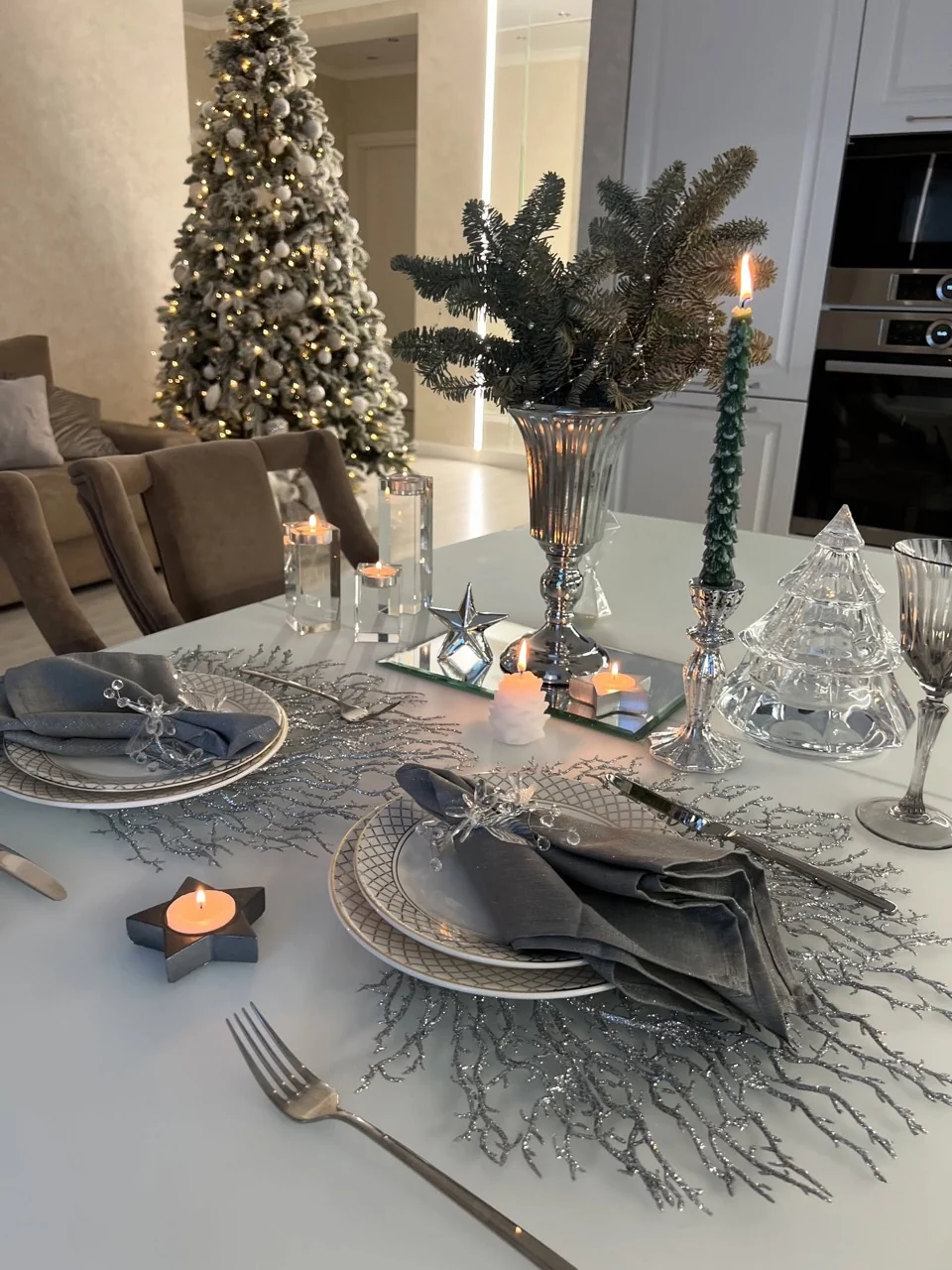 Christmas table set with grey napkins, silver branch-shaped placemats, evergreen stems and lit candles.