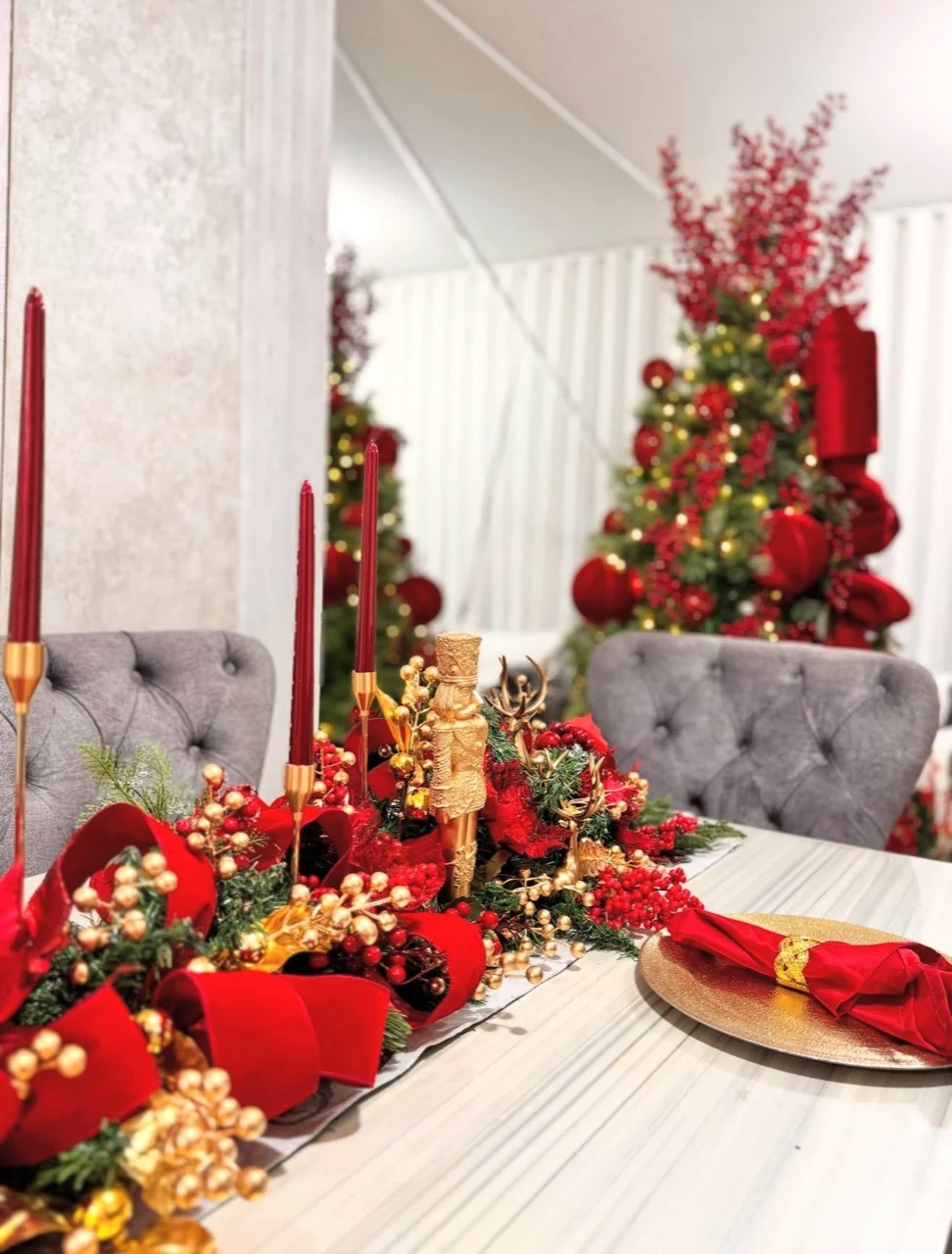 Red and gold Christmas table runner with ribbon, berries, a nutcracker and tall red candles, with a decorated tree behind.