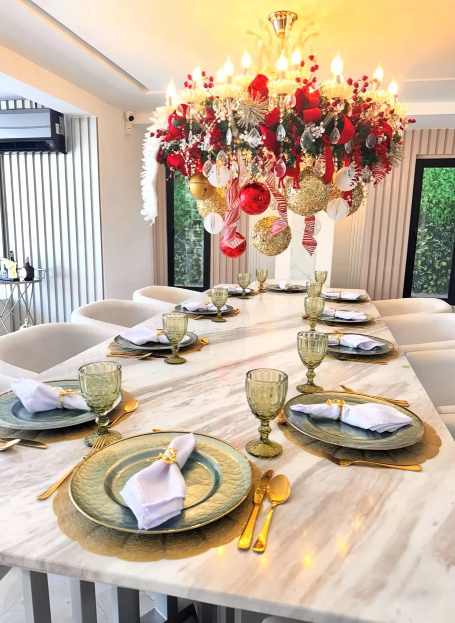 Long dining table with gold chargers and green goblets beneath a chandelier decorated with greenery, ribbon and red and gold baubles.