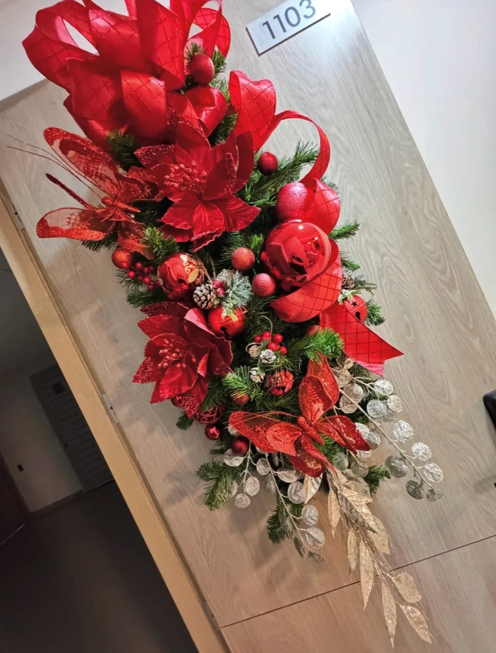 Vertical Christmas door swag with red poinsettias, ribbon, baubles, pinecones and trailing gold foliage.
