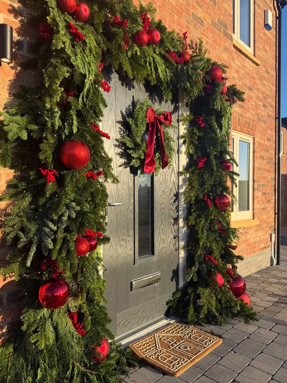 Dark front door framed by thick evergreen garlands with red baubles and berries, with a red-bow wreath in afternoon sunlight.
