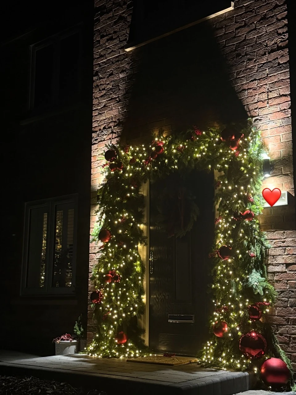 Outdoor Christmas doorway at night with warm fairy lights woven through evergreen garlands and red ornaments.