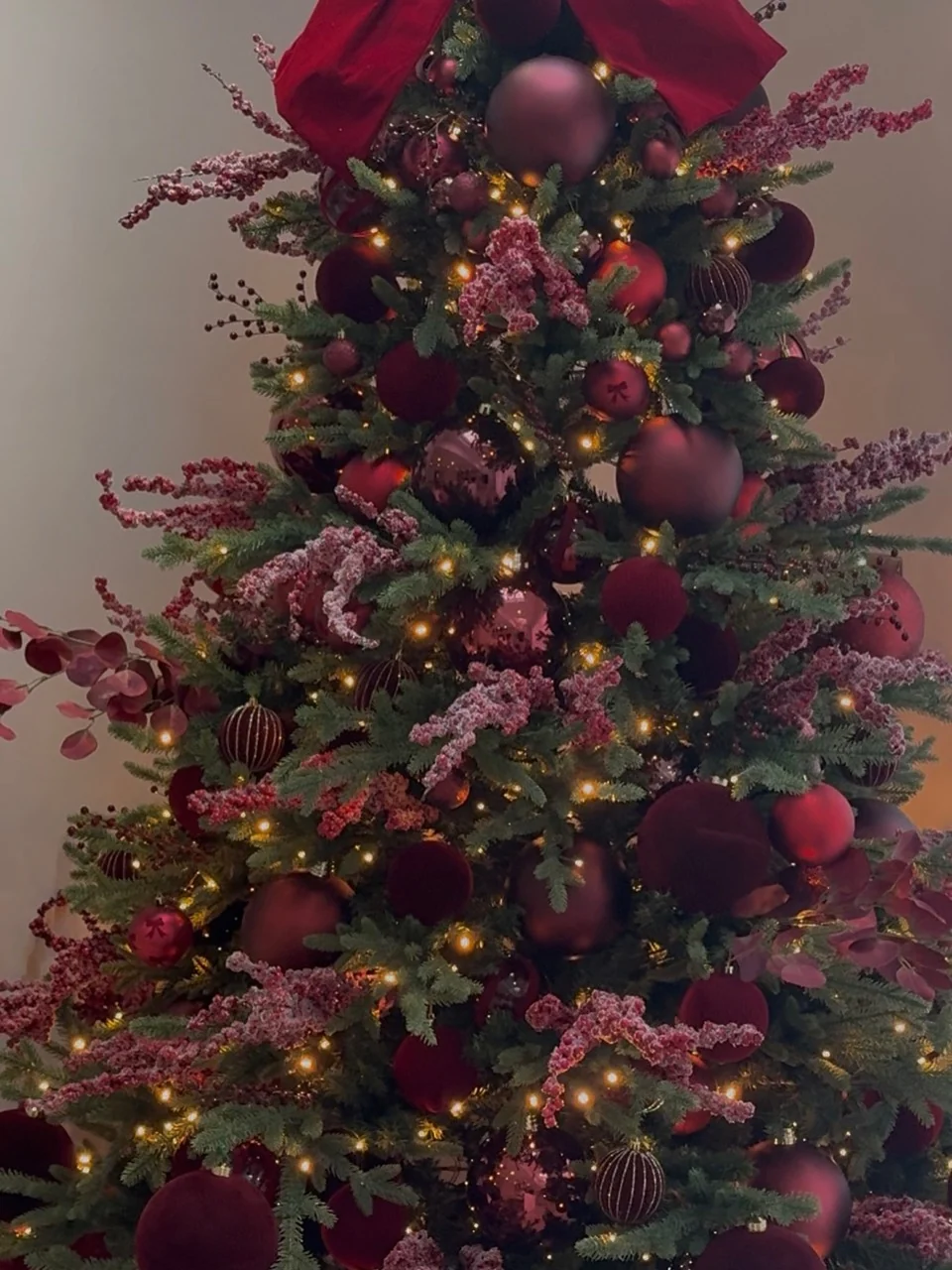 Christmas tree decorated with burgundy velvet and shiny baubles, frosted berry sprays, warm lights and a large burgundy bow.