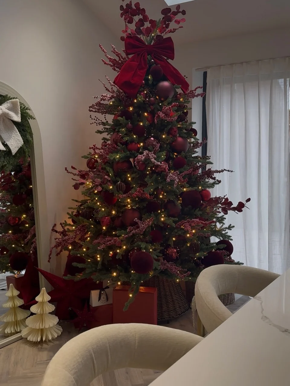 Burgundy Christmas tree with a large velvet bow, berry sprays and warm lights in a woven basket beside a mirror and cream dining chairs.