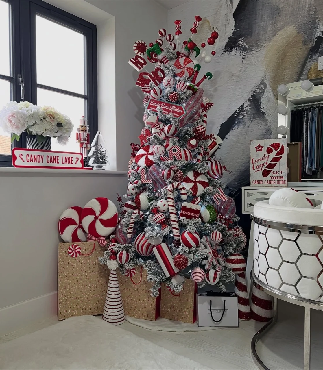 Flocked Christmas tree filled with red and white peppermint ornaments, striped ribbon and candy-cane decorations beside wrapped gifts.
