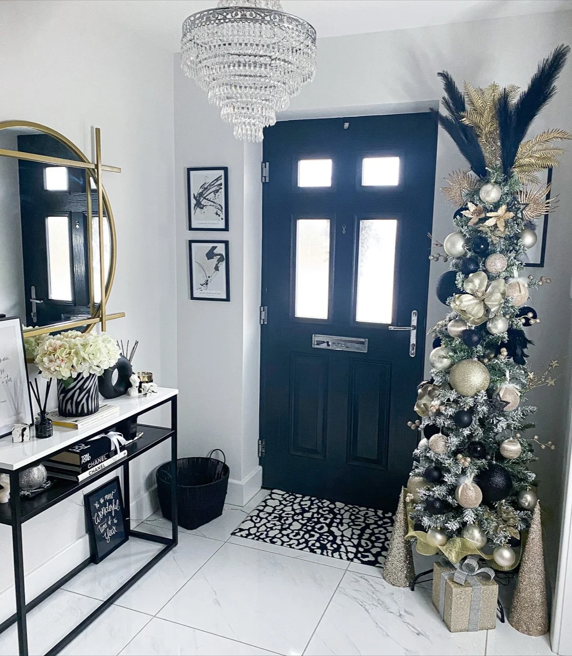 Slender Christmas tree decorated with black and gold ornaments, metallic foliage and feathers beside a dark front door and gold-framed mirror.